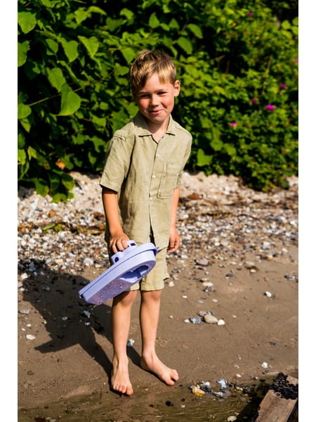 Person ved vandkant med blå legebåd i vandkanten mellem sten og sand.
